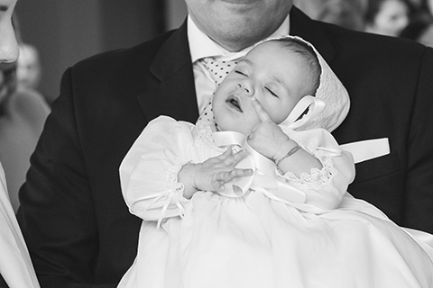Imagen en blanco y negro de un bebé sobre los brazos su padre, está dormido con un dedo sobre su mejilla mientras se celebra la ceremonia, la imagen la realizó un fotógrafo de bautizo en Madrid.