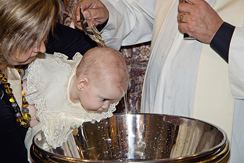 Bebé recibe el agua bautismal en una iglesia, se ve a la niña en los brazos de su abuela, está mirando hacía la pila y el agua cubre su cabeza, una fotografía de bautismo en Madrid.