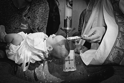 Mano de un cura con una concha en su mano llena de agua, el bebé en brazos espera el bautismo por parte del cura en un reportaje de bautizo en la Ermita de la Moraleja.