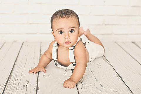 Fotografía de bebé de 9 meses en fondo blanco en estudio fotográfico. El niño esta tumbado boca abajo con la cabeza levantada, lleva un peto de color blanco.
