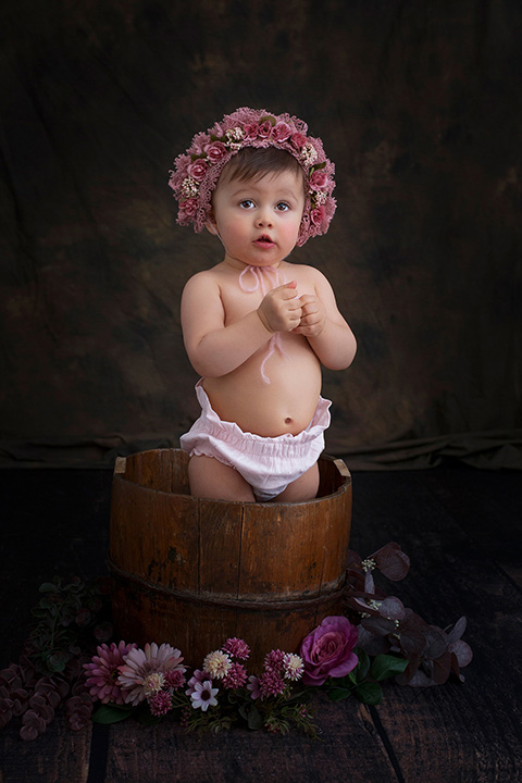Sesión fotográfica de bebé con una niña en un cubo y con un gorro de flores, simulando que ella misma es una maceta con flores rosas.