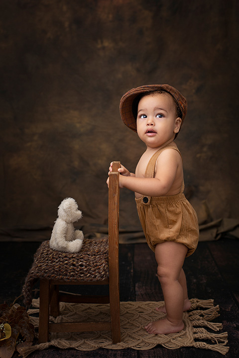 Bebé con boina y pelele con tirantes, de pie sujetado a una silla y con un oso sentado en sus sesiones de fotos de bebé