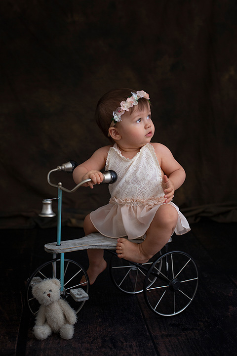 Niña en una bicicleta con una corona de flores, jugando con un oso mientras le hacen unas fotos de bebé.