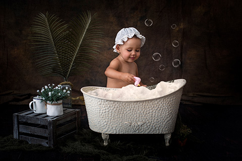 Reportaje de bebé, niña con gorro sentada en una en una bañera, jugando con la espuma y un pato de juguete.