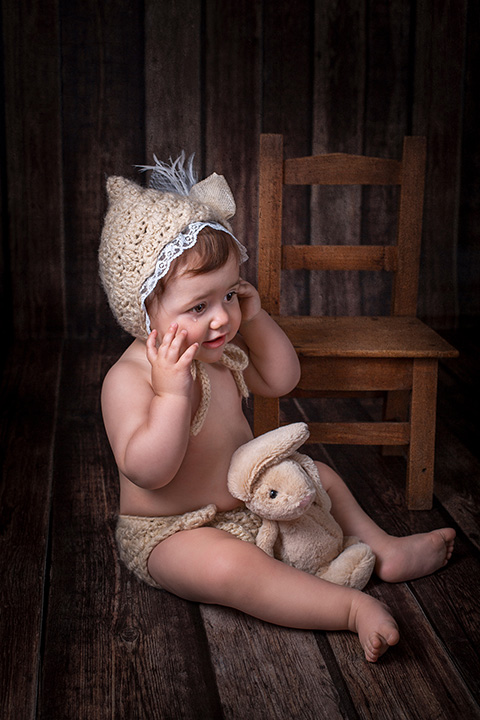 Foto de una niña sentada en el suelo con una capota y un conejo, tiene las manos sobre la cara y una lágrima le cae sobre su mejilla, posa mientras el fotógrafo de bebés captura imágenes.