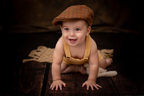 Book de fotos de bebé en estudio de fotografía. Niño de un año tumbado boca abajo, apoyado con manos y pies. Lleva un peto y una boina de color tostado.