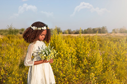 Niña de pelo rizado con un vestido blanco de encaje sobre un campo de flores amarillas, sujeta un ramo a juego en su sesión de primera comunión en exterior.