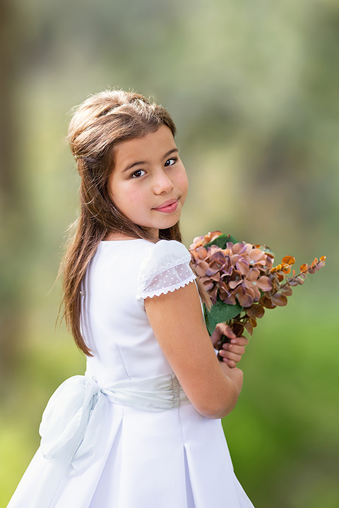 Fotógrafos de comunión realizan las fotos de una niña en exteriores, con un vestido blanco y unas flores entre sus manos, está de espaldas y gira su cara hacía la cámara.