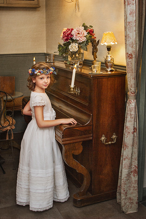 Niña con un vestido blanco y corona de flores tocando el piano, imagen realizada por un fotógrafo profesional de comunión.