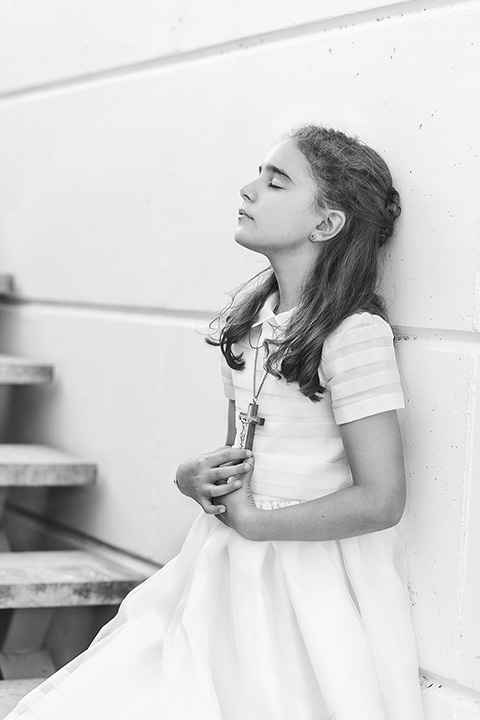 Sesión de fotos de primera comunión en blanco y negro de una niña apoyada sobre una pared, tiene los ojos cerrados y sus manos están tocando una cruz de madera.