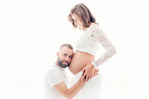 hombre vestido de blanco apoyado sobre la barriga de su mujer que le mira, escuchando los latidos de su futuro bebé mientras les toman unas fotografías de maternidad.