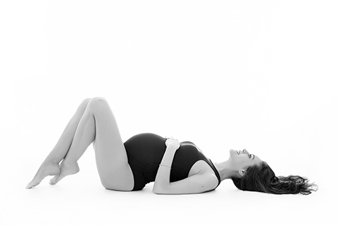 Mujer tumbada en el suelo boca arriba, lleva un body negro, tiene las piernas dobladas y se está abrazando la tripa en una sesión fotográfica para embarazas en estudio.