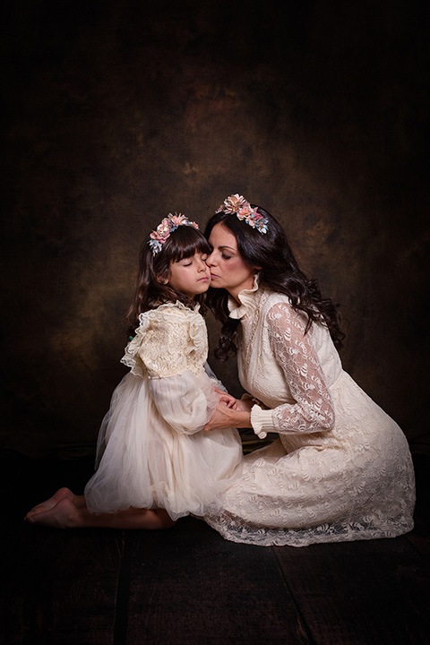 Madre besa a su hija en la mejilla, están de rodillas sobre un fondo de estudio color marrón, llevan un vestido beige de encaje y una corona de flores en su cabeza en una sesión de fotos fin art de familia.