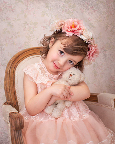 Foto de medio plano de una niña sentada abrazando un oso de peluche, lleva una corona de flores y un vestido rosa de encaje realizada por un fotógrafo infantil en Madrid.