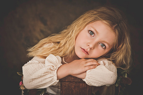 Primer plano de una niña rubia, con ojos azules, está apoyando su cara sobre sus manos realizada por fotógrafos infantiles en Madrid.