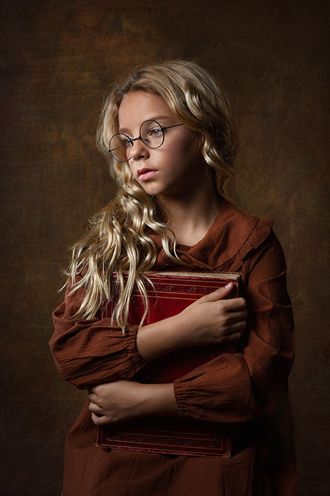 Sesiones de fotos infantiles en Madrid de una niña rubia con pelo rizado y gafas, lleva un vestido marrón y está sujetando un libro antiguo entre sus manos.