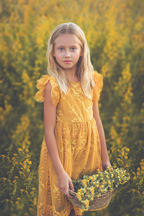 Niña rubia con ojos azules en un campo de flores amarillas con un vestido a juego, está sujetando una cesta con las flores en una sesión de fotos infantil en exteriores.