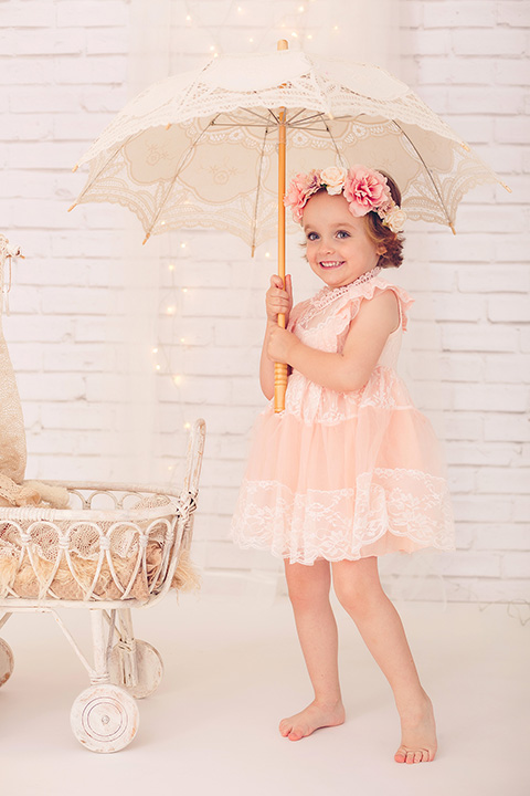 Book de fotos profesional infantil, niña con vestido rosa y corona de flores con paraguas, el fondo decorado con un cochecito de mimbre.