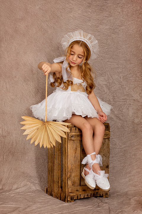 Fotógrafo de moda infantil, niña vestida de blanco con capota sentada sobre un cajón de madera, está sujetando un paipay con su mano.