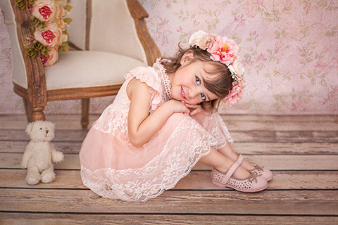 Fotografía para catálogos de moda infantil, niña sentada en el suelo con un vestido y zapatos rosas, con corona de juego en la cabeza, está con las rodillas flexionadas, con las manos y su cara apoyada sobre ellas.