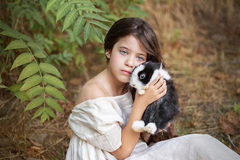 Imagen de una niña con ojos azules en el campo, lleva un vestido y tiene entre sus manos un conejo blanco y negro, por detrás de ella hay unas ramas verdes, en una sesión de fotos infantil fin art en exterior.