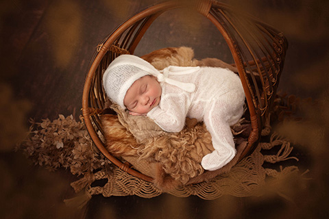 Bebé dormido y tumbado boca abajo en una cesta de ratán, está sobre una manta de pelo y va vestido con un gorro y pijama blanco.