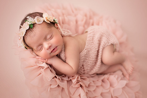 Niña recién nacida tumbada sobre una manta rosa con forma de flor, esta dormida de lado con su mano apoyada en su mejilla, además lleva una corona de flores en su cabeza.