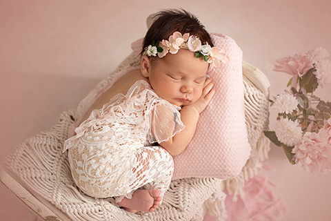 Bebé recién nacido con 10 días de vida, tumbada en una cama boca abajo, dormida sobre un cojín en tonos rosas, viste un body en color blanco.