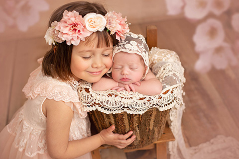 Imagen de un niña con su hermana recién nacida, la bebé esta en un cubo de troncos de árbol que la mayor sujeta con sus manos.
