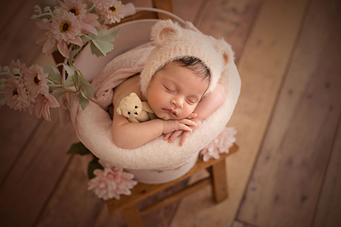 Fotografía newborn vintage de una niña con días de vida, dormida entro de un cubo rosa sobre una silla de madera, lleva un gorro con forma de oso y sujeta un oso beige en su brazo derecho.