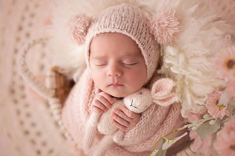 Niña recién nacida en una sesión de fotos, dormida sobre una cesta de mimbre, lleva un gorro rosa con pompones a los lados y sujeta con sus manitas un conejo del mismo color.