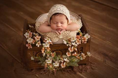 Niña con tan solo 7 días de vida con gorro de encaje, está dormida sobre un cubo beige con flores alrededor.