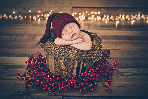 Bebé metido en un cubo de madera rústico, lleva un gorro granate de lana, su cara está apoyada sobre sus manos y está dormido, alrededor tiene acebo artificial y al fondo se ven lucecitas, imagen realizada en un reportaje de recién nacido de Navidad.