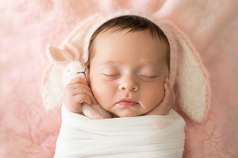 Bebé niña con un gorro de lana con orejas de conejo en color rosa y blanco, está dormida y envuelta en una tela de color blanco, solo se ven sus manos que apoyan sobre sus mejillas y una de ellas sujeta un conejo de lana rosa en una fotografía de newborn en Madrid.