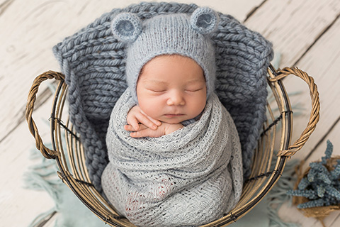 Bebé dormido en un cesto, con el cuerpo envuelto en una tela, solo se ven sus manos y su carita que apoya sobre ellas, lleva un gorro de lana azul con orejas de oso en una sesión newborn con atrezzo y decorado.