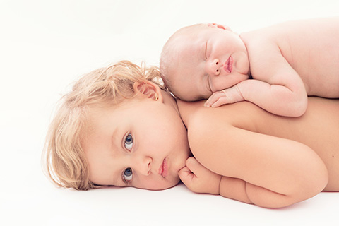 Niña tumbada boca abajo, apoyada su mejilla en el suelo, tiene a su hermana bebé durmiendo en su espalda, las dos están sin camiseta, es una imagen realizada por una fotógrafa especializada en sesiones de recién nacido.