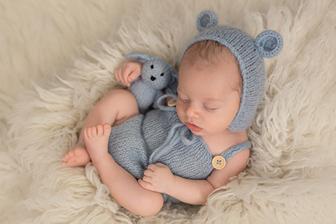 Bebé dormido en una manta de pelo blanca, lleva un gorro de lana con orejas de oso en tono azul y un body a juego, sujeta con una de sus manos un conejo de lana, en un reportaje de fotos de recién nacido en Madrid.