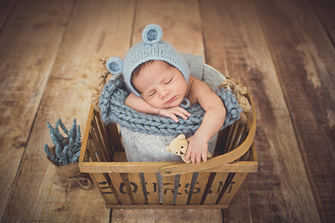 Bebé dormido metido en un cubo de metal sobre otra cesta de madera, el niño tiene su cabeza apoyada en uno de sus brazos y la otra cuelga con un oso en la mano, lleva un gorro de lana de oso en color azul, imagen realizada en un reportaje de recién nacido con atrezzo y vestuario.