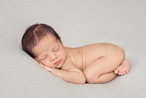Imagen de recién nacido en un puff, sesión newborn en posición boca abajo y con las piernas cruzadas y las manos debajo de su mejilla sobre un fondo gris.