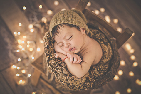 Bebé de 9 días dormido, está dentro de un cubo rústico que está sobre una silla de madera, sus manos están apoyadas sobre el cubo y su cara sobre ellas, alrededor hay muchas luces dado que es una sesión de fotos de recién nacido navideña.