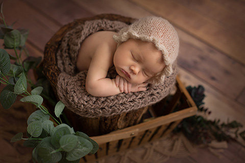 Bebé dormida en un cubo de madera rústico dentro de una cesta de madera, la niña lleva una capota de lana beige y su cara apoya sobre sus manos, realizada por fotógrafos newborn Madrid.