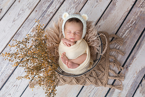 Niño dormido en una cesta, está envuelto en una tela de color amarillo pastel, lleva un gorro de pelo con orejas de oso, a través de la manta asoman sus manos y pies, imagen realizada por fotógrafos profesionales de recién nacido.