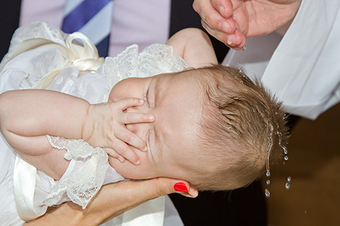 Bebé en un reportaje de bautizo en Madrid, justo está recibiendo el agua bautismal sobre su cabeza, tiene la manita en la cara, sobre la imagen también aparece una parte de la mano del cura. 