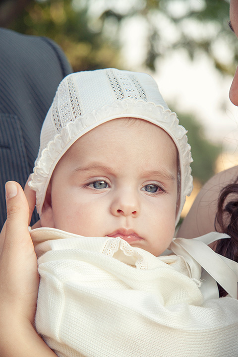 Reportajes fotográficos de bautizo realizados en exteriores de un bebé sobre los brazos de su madre, lleva una capota y un traje en color beige.