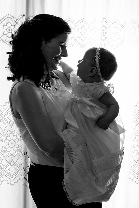 Imagen en blanco y negro de una madre y su hija, la bebé está en brazos y las dos se están mirando, fotografía de bautizo en el domicilio tomada a contra luz de una ventana.