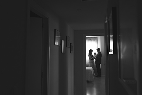 Imagen en blanco y negro de unos padres con su bebé, están a contra luz de una ventana y se ve que están al fondo de un pasillo en una fotografía de bautizo en el domicilio.
