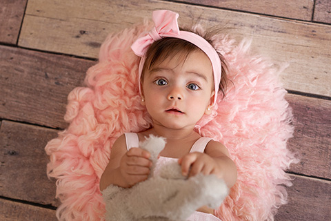Fotografía de bebé niña con ojos azules, tumbada en el suelo con peluche y lazo rosa. 