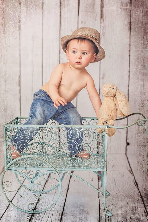 Niño rubio con sombrero, jugando en un carro con un conejo de peluche, capturada por un fotógrafo de bebés.