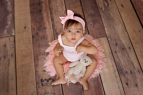 Niña con un oso, con un peto rosa y lazo en la cabeza sobre una manta y suelo de madera, tomada por fotógrafos profesionales de bebés.