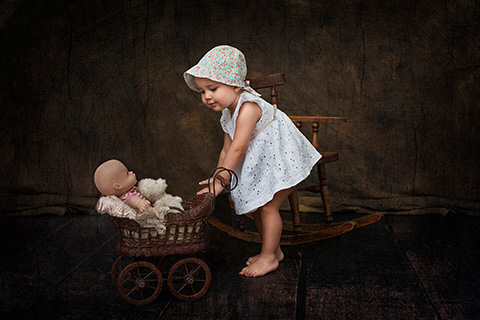 Reportaje de bebé, niña con vestido y sombrero, lleva un carro con un muñeco de bebé y un oso.